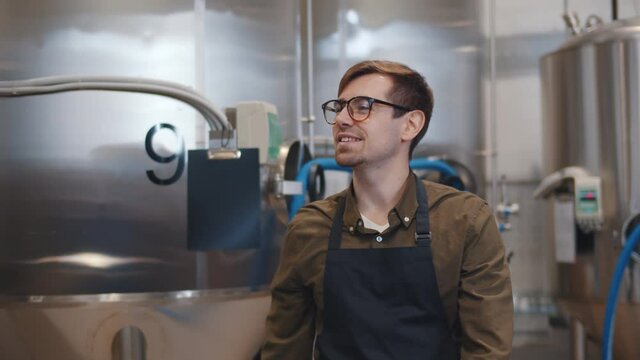 Young brewer in glasses and apron walking near metal tanks at factory