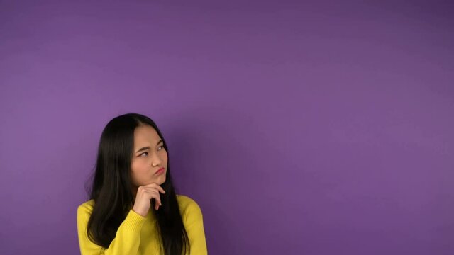 puzzled young asian woman in bright yellow sweater looks right. Free space for text and speech bubble. isolated purple background. 4K