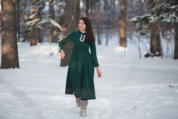Friendship of an owl and a girl. Myth, fairy tale. Mystical spirit. Fantasy female elf walks with an owl in the winter forest. Beautiful brunette brunette girl in a green vintage dress. Tame bird.