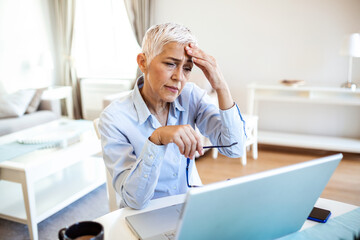 Senior frustrated woman working from home office in front of laptop, having a headache.Mature woman...
