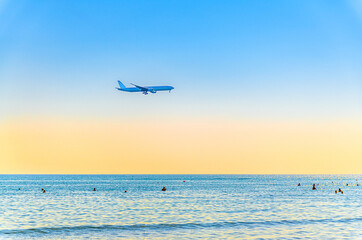 Fototapeta na wymiar Airplane flying low above sea and people tourists swimming in water, clear blue orange sky at sunset, plane preparing to land at Larnaca airport above Mediterranean sea