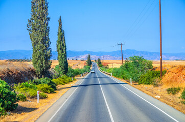 Fototapeta premium Landscape of Cyprus with cars vehicles riding asphalt road in valley with yellow dry fields, cypress trees and roadside poles, Troodos mountain range and hills, clear blue sky in sunny day background