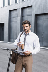 bearded businessman in suit holding coffee to go and using smartphone near e-scooter