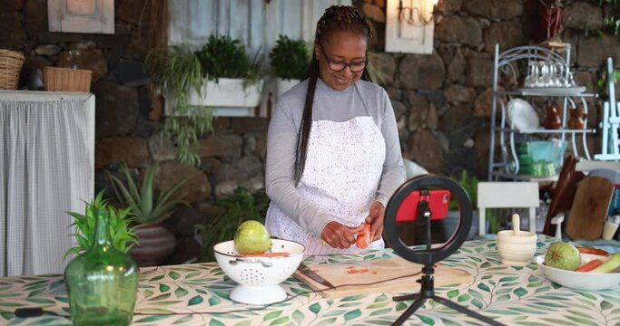 African senior woman cooking outdoor while streaming online for webinar masterclass lesson at home - Food and influencer concept 