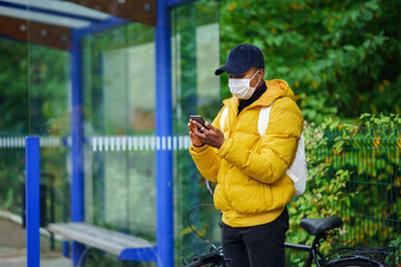 Young man with face mask on bus stop outdoors in town, using smertphone. Coronavirus concept.