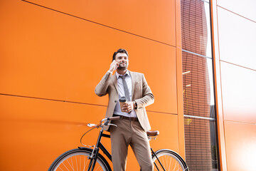 smiling businessman talking on smartphone and holding paper cup near bicycle and building with orange walls