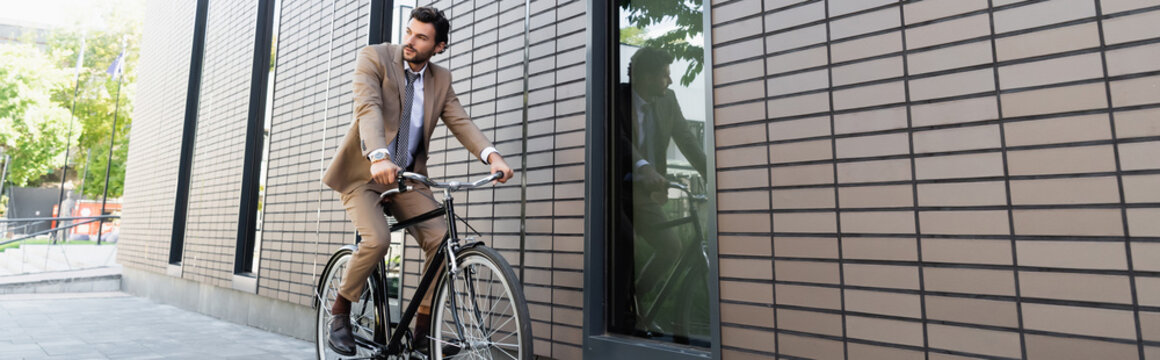 bearded businessman in suit riding bicycle and looking away near building, banner
