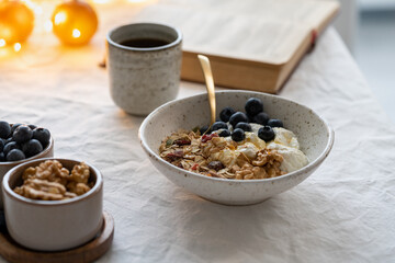 Reading book and eating healthy Christmas holiday winter breakfast with granola muesli and yogurt in bowl on white table background, lights garlands in bokeh. Organic morning diet meal with oat 