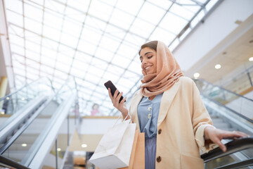 Low angle portrait of smiling Middle-Eastern woman looking at smartphone screen while standing on escalator in shopping mall, copy space