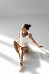 graceful african american ballerina in dress sitting on floor on white background