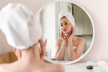 A young woman in white clothes and with a towel on her head looks in the mirror and applies gold plasters under her eyes, taking care of her skin in the morning. Woman applying gold spots on her face.