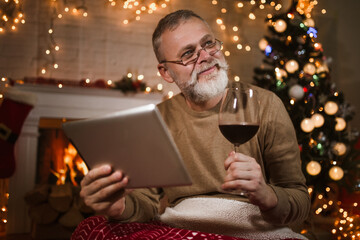 Man drinking wine from glass and using digital tablet sitting and relaxation near fireplace and christmas tree.