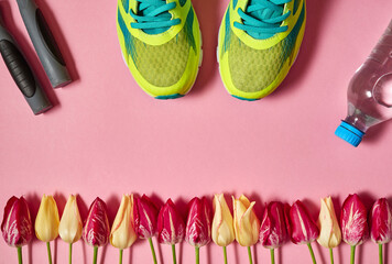 Pair of sport shoes, skipping rope, bottle of water and tulips on pink background. New sneakers, copy space. Overhead shot of running foot wear. Top view, flat lay