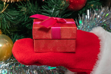 Close up shot of Santa Claus holding a red gift box in one hand. Christmas tree with red and golden balls, white tinsel blurred in the background. New Years, Christmas, winter holidays concept