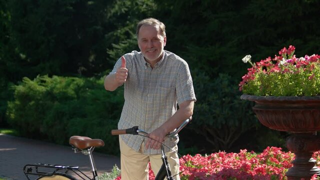 Smiling mature man giving thumb up outdoors. Attractive senior man on bicycle at park on a sunny day. Active lifestyle concept.