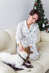 Happy young woman wearing pajama with lovely dog in living room with christmas tree. Holidays concept.