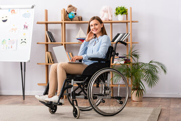 Full length of smiling psychologist with laptop looking at camera while sitting in wheelchair in office