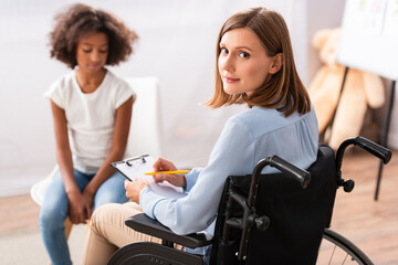 Positive psychologist writing on clipboard while sitting in wheelchair with blurred african american girl on background