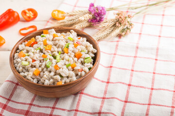 Pearl barley porridge with vegetables in wooden bowl on a white wooden background and linen textile. Side view, selective focus.