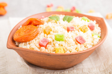Bulgur porridge with dried apricots and candied fruits in clay bowl on a gray concrete background and linen textile. Side view, selective focus.