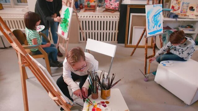A lesson in an art school, children sit at easels, paint with paints, a teacher helps students cope with their creative tasks. Child with special needs at a painting workshop.