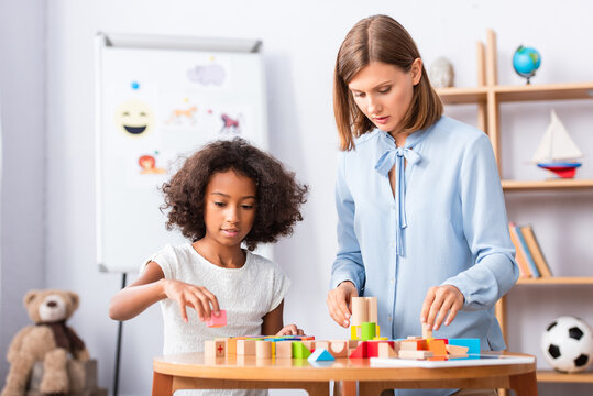 Psychologist talking to african american girl playing with colorful wooden blocks on coffee table on blurred background