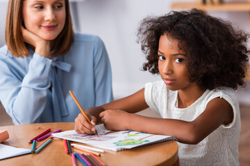 Obraz premium African american girl looking at camera while drawing with colored pencils near positive psychologist on blurred background