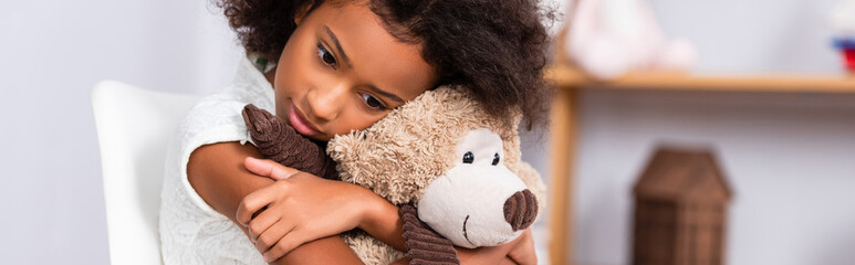 Depressed african american girl hugging teddy bear with blurred office on background, banner
