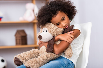 Depressed african american girl with autism hugging teddy bear while sitting on chair with blurred office on background