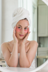 Young lady enjoying morning anti age anti wrinkle routine in bathroom after showering. Headshot smiling pretty woman wearing towel applying hydrogel patches on under eye area, looking at mirror.