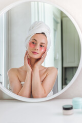 Young lady enjoying morning anti age anti wrinkle routine in bathroom after showering. Headshot smiling pretty woman wearing towel applying hydrogel patches on under eye area, looking at mirror.