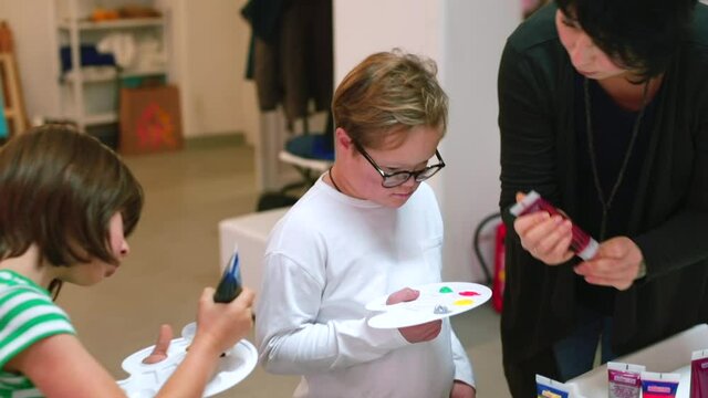 The beginning of the lesson at art school, children put paint on a palette. A child with Down syndrome is assisted by a teacher.