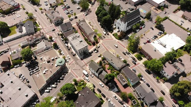 A daytime birds-eye drone view of a suburban downtown traffic grid among the buildings and shops, cars and other vehicles driving on the roadway.