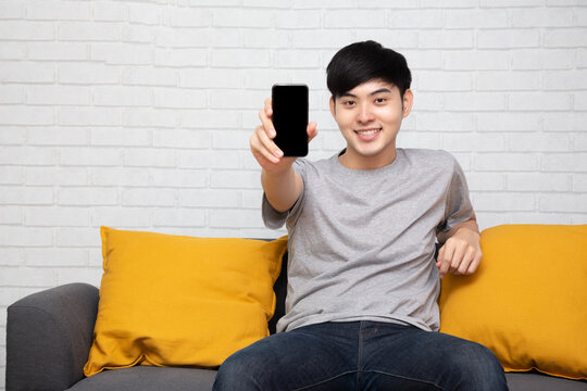 Young Asian Man Sitting On Sofa And Showing Present Mobile Phone Application At Home