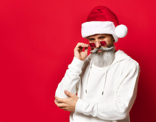 Fototapeta na wymiar Winter fashion and sales. Portrait of senior handsome man in santa hat winking standing on red background. Hipster holds his glasses and looks at the camera. Place for text.