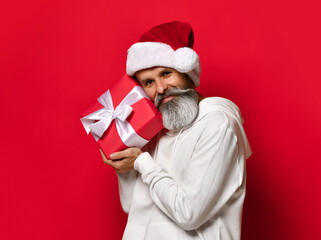 Handsome senior bearded man holding a gift box over red background and looking at the camera. Santa Claus wishes a Merry Christmas and a Happy New Year.