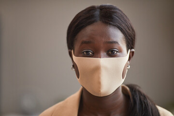 Minimal front view portrait of young African-American woman wearing mask in office and looking at camera, copy space