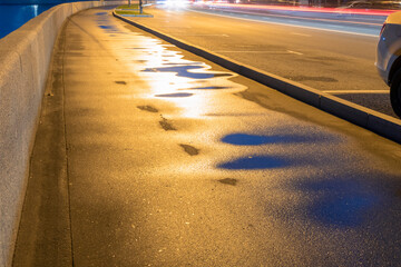 Empty wet sidewalk with rain puddles, road and side road parking lots. Coming cars lighten area....