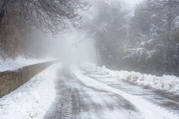 Fototapeta premium Icy mountain road through a snowy forest shrouded in thick fog on a winter day. Dangerous driving conditions.