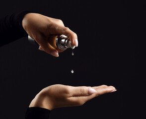 Hands of woman pouring moistirizing oil for hair rehabilitation or treatment from bottle to hand before procedure over black background, close-up. Haircare, beauty, wellness, cosmetics concept
