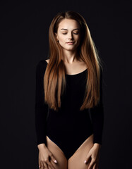 Young beautiful woman with long silky straight hair in black body with eyes closed standing and posing over dark background. Haircare, beauty, wellness concept