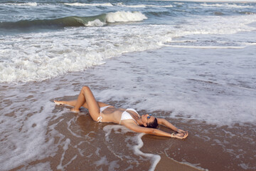 young sexy woman in a white swimsuit and straw hat is lying on a sandy beach by the sea in the summer on vacation