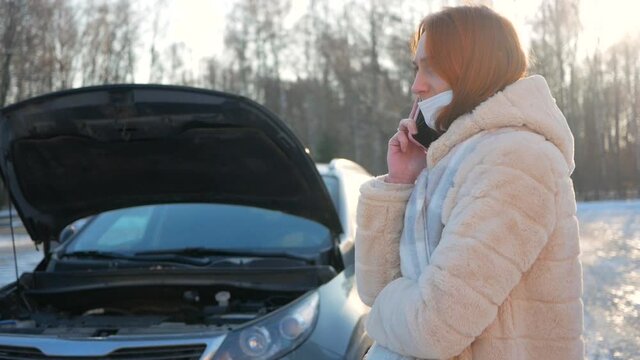 A young girl in a medical mask had a car breakdown in winter. Calls technical assistance on the road. Dangerous situation on the winter road