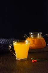 Hot tea with sea buckthorn in a glass pot on the dark wooden table, knitting blanket near. Healthy drink, cozy winter night. Vertical orientation, copy space.