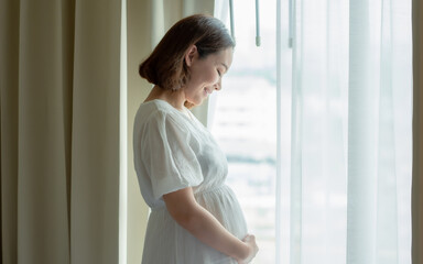 Asian pregnant woman wearing white dress and standing beside window