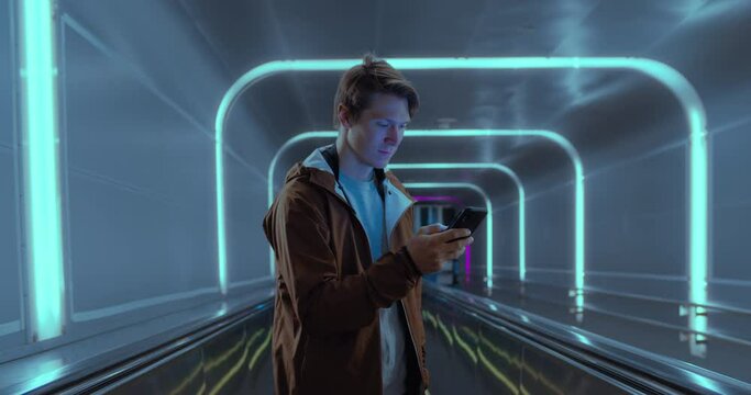 Portrait of modern urban millennial man scroll news or social media on smartphone. Generation z hipster man stand in futuristic cool scenery. Neon lights and new normal 2020 setup. Cinematic youth