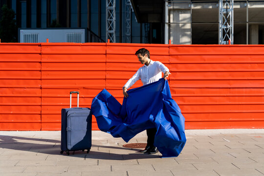 Businessman swinging blue cloth next to wheeled luggage on pavement in the city