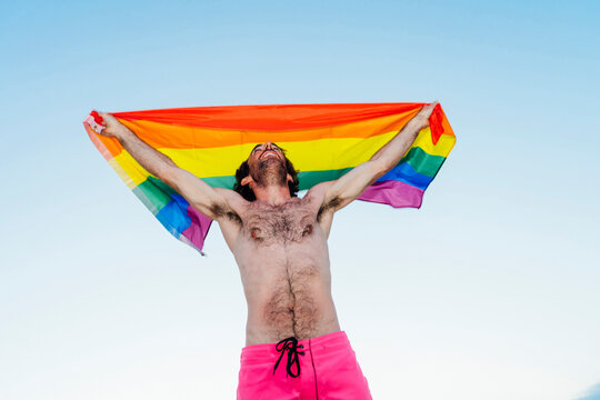 Laughing man holding gay pride flag against clear sky
