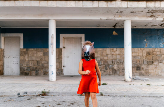 Little girl with a horse's head and a red dress walking away from an abandoned house