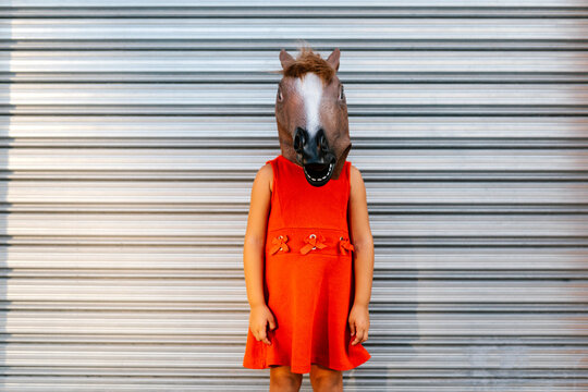 Litle girl with a horse's head and a red dress in front of  metal shutter